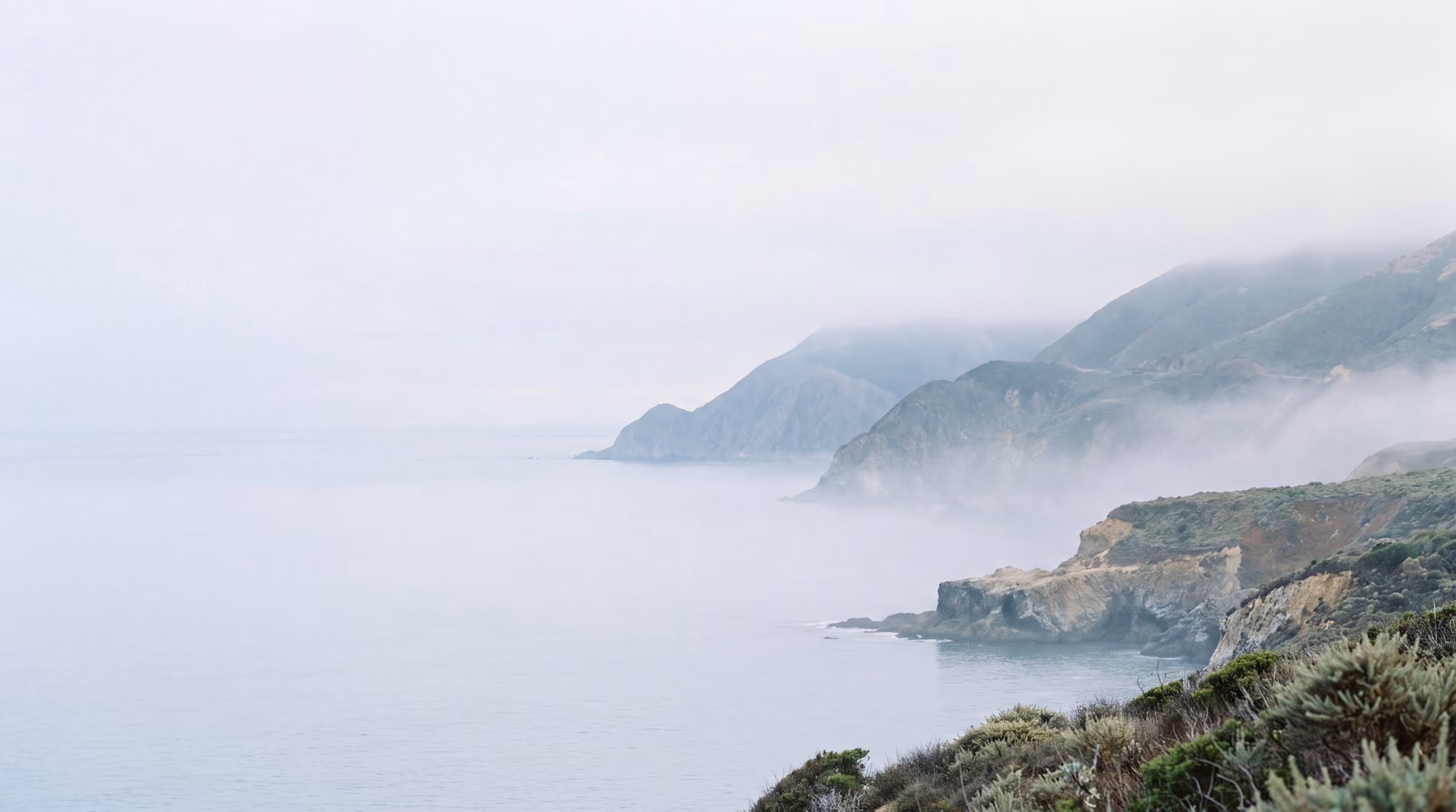 Headlands along a calm coast in soft morning fog, the sea still and pale